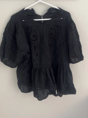 Svart blus med broderade detaljer - Svart blus från zara med broderade och utskurna detaljer framtill. Blusen har en lös passform, volang nertill och korta ärmar. Materialet ser ut att vara tunn bomull eller liknande, vilket ger en luftig känsla. Perfekt för en trendig och avslappnad look.