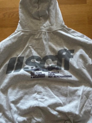 Grå hoodie med tryck från Scuffers - Snygg grå hoodie med stor svart text och grafiskt tryck på ryggen. Hoodien har luva och ribbade muddar vid ärmslut och nederkant. Perfekt för dig som gillar streetwear och vill ha en chill look.