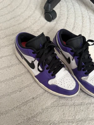 Nike Air Jordan 1 Low Court Purple - Säljer ett par Nike Air Jordan 1 Low Court Purple sneakers med vit och lila ovandel, svarta detaljer och svart swoosh. Skorna har svart plös och svarta skosnören. Klassisk låg siluett och tillverkade i läder för en clean och stilren look.