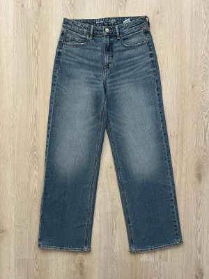 Blå raka jeans med hög midja - Säljer ett par klassiska blå jeans med rak passform och hög midja. Jeansen har fem fickor, knappgylf och subtila slitningar framtill för en vintagekänsla. Tillverkade i kraftigt denimtyg som ger en tidlös look.