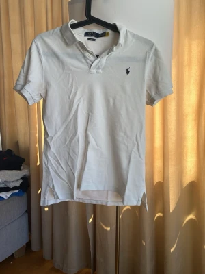 Vit pikétröja från Polo Ralph Lauren - Klassisk vit pikétröja från Polo Ralph Lauren med korta ärmar och krage. Tröjan har knappar vid halsen och den ikoniska loggan broderad på bröstet. Tillverkad i mjuk bomull för en bekväm känsla.
