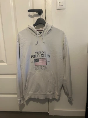 Vit hoodie Vinson Polo Club - Vit hoodie från Vinson Polo Club med tryck av amerikansk flagga och text på bröstet. Klassisk känguruficka framtill, dragsko i huvan och ribbade muddar. Skriv till mig om funderingar och pris!!