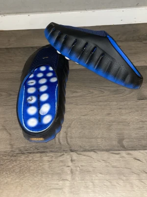 Navy tofflor med mönster Nike mind 001 - Säljer ett par unika tofflor i blått och svart med sportig design. Ovandelen har ett prickigt textilmönster och sulan är kraftig med räfflad kant. Innersulan är blå med vita cirklar och text. Perfekta för att chilla hemma eller på gymmet. Nike mind 001