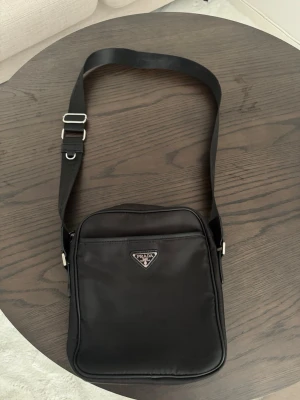Prada crossbody väska  - Säljer en Prada-väska i mycket fint skick som jag fått i gåva. Har aldrig använt den. Väskan har Prada-märkning, QR-kod och fina silverfärgade metalldetaljer. Jag har inget kvitto eller äkthetsintyg och kan därför inte garantera äktheten. Väskan har däremot visats för Stockholms Auktionsverk och de bedömde att väskan är äkta.