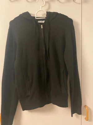 Svart stickad hoodie med dragkedja - En svart stickad hoodie med dragkedja framtill och två fickor. Tröjan har långa ärmar, ribbade muddar och en klassisk huva. Perfekt för lager på lager och enkel att matcha med olika outfits.