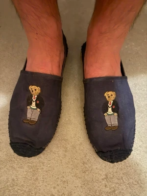 Polo Ralph Lauren mörkblå espandrillos med polobjörnen - Säljer ett par mörkblå espandrillos från Polo Ralph Lauren med broderad nallebjörn på ovansidan. Nallen har kavaj, slips och byxor vilket ger en preppy look. Tofflorna har rundad tå och mjuk sula, perfekta för sommaren!.