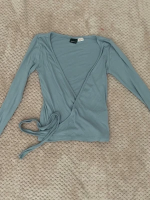 Ljusblå omlottopp från Gina Tricot - Säljer en ljusblå omlottopp från Gina Tricot i mjukt material. Toppen har långa ärmar och knytband i midjan för en snygg passform. Perfekt att styla med jeans eller kjol för en trendig look.