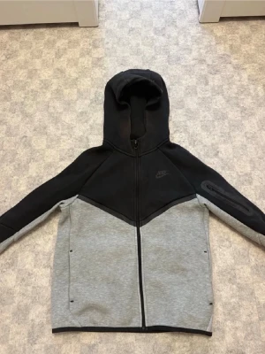 Nike Tech Fleece hoodie svart/grå - Nike Tech Fleece hoodie i svart och ljusgrått med dragkedja framtill och huva. Snygg tvåfärgad design, diskret Nike-logga på bröstet och ärmen. Mjukt och lätt material som ger en modern look. Perfekt för en avslappnad streetstyle.