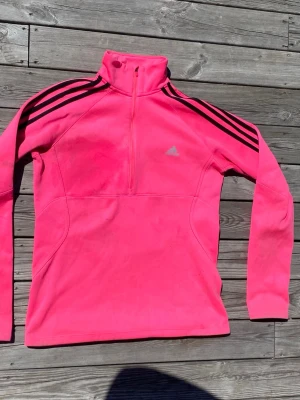Rosa fleece-tröja från Adidas - Snygg rosa fleece-tröja från Adidas med svart detalj i form av tre ränder på axlarna. Tröjan har half zip, hög krage och lång ärm. Perfekt för träning eller som ett extra lager under kalla dagar.