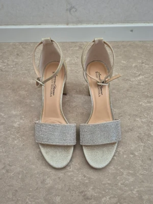 Platåsandaletter - Säljer ett par glittriga sandaler med blockklack från Badgley Mischka med breda remmar täckta av små stenar och justerbara ankelband. Skorna har öppen tå och stabil klack, perfekta för att lyfta vilken outfit som helst. Stl 8 är amerikansk storlek så det blir stl 38/39. Använt endast 1 till 2 timmar.