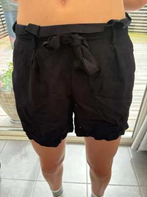 Svarta shorts med knytband - Modellen är 157. Säljer ett par svarta shorts med hög midja och ett snyggt knytband i midjan. Shortsen har en avslappnad passform och lätt papperspåseffekt upptill. Perfekta för varma dagar och enkla att matcha med olika toppar.
