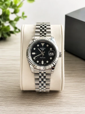 Seiko automatisk klocka i silver - Stilren Seiko automatisk klocka med svart urtavla, tydliga index och datumvisning. Klockan har en klassisk länk i silverfärgat rostfritt stål och en räfflad bezel. Baksidan är transparent så du ser urverket. Perfekt accessoar för dig som gillar tidlös design.