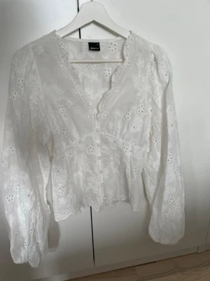 Vit broderad blus från Gina Tricot - Säljer en vit blus från Gina Tricot med broderade blommor och hålmönster. Blusen har v-ringning, knappar framtill och långa puffärmar. Tillverkad i ett tunt, luftigt tyg som ger en romantisk vibe. Perfekt att styla med jeans eller kjol.