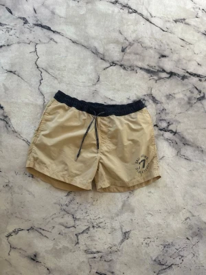 Beige badbyxor Sail Racing - Snygga beige badbyxor från Sail Racing med svart resårmidja och dragsko. Tryck med texten 'Sail Racing Doubleseven 77' på vänster ben. Perfekta för stranden eller poolen, tillverkade i snabbtorkande material.