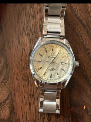 Omega Seamaster silver herrklocka - Säljer en stilren Omega Seamaster herrklocka med silverfärgat armband i metall och ljus urtavla med diskreta ränder. Klockan har datumvisning och tydliga markeringar. Extralänkar medkommer givetvis!