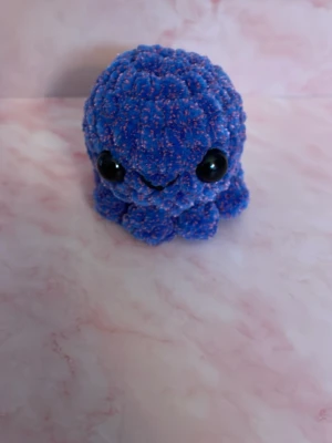 Blå virkad bläckfisk plushie - Söt handgjord bläckfisk plushie i blått garn med inslag av rosa och stora svarta ögon. Perfekt som dekoration eller mysig maskot. Mjuk och fluffig känsla tack vare det virkade materialet. Höjd 6,5 cm