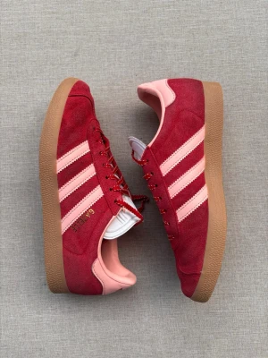 Adidas Gazelle röda sneakers - Säljer ett par Adidas Gazelle sneakers i rött mocka med ljusrosa detaljer och klassiska tre ränder på sidan. Skorna har gummisula i brunt och röda skosnören med vita inslag. Perfekt för dig som vill ha en retro och färgstark look.