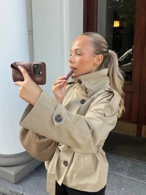 Kort trenchcoat - Gina Tricot  - Säljer en superfin beige trenchcoat från Gina Tricot i storlek S. Endast använd vid två tillfällen och i mycket fint skick. Modellen är slutsåld online.                                                                 Kappan har en stilren hög krage, ett skärp som kan knytas både fram och bak för önskad passform samt ett rejält tyg med fin kvalitet som gör den perfekt för både vår och höst.         Nypris: 699 kr
