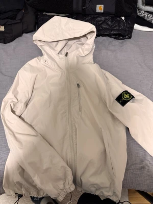 Beige vindjacka från Stone Island - Snygg beige vindjacka från Stone Island med huva och dragkedja framtill. Jackan har en bröstficka med dragkedja, två sidofickor och den klassiska Stone Island-loggan på vänster ärm. Tillverkad i lätt och slitstarkt material, perfekt för blåsiga dagar.