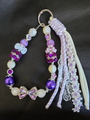 Handgjort lila och vitt pärlprydd accessoar med rosetter - Handgjord accessoar med lila, vita och glittriga pärlor, dekorerad med flera rosetter och detaljer i silver. Hängande band i spets och flätad textil ger en extra söt touch. Perfekt som smycke till väskan eller nyckelringen.