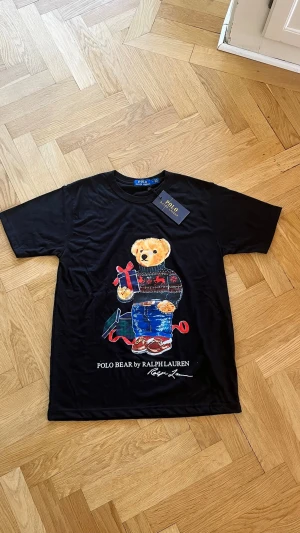 Svart Polo Bear t-shirt från Ralph Lauren - Svart t-shirt från Ralph Lauren med ikoniska Polo Bear-trycket framtill. Björnen har en julig tröja och håller i ett paket. Rund halsringning och klassisk passform. Perfekt statement-plagg för dig som gillar lekfulla detaljer. Denna t shirten har måtten = Axel till axel 43cm och Axel ner till botten 65cm. Jag försöker att skicka samma dag skriv till mig om ni vill göra ett bundle med den andra t shirten som ligger upp fast i M och vit👍😊