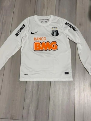Vit långärmad Santos FC matchtröja - Säljer en vit långärmad matchtröja från Santos FC med tryckta sponsorer och klubbmärke på bröstet. Tröjan har Dri-FIT-material för bra ventilation och komfort. Svarta detaljer och orange BMG-logga framtill. Perfekt för fotbollsträning eller som supporterplagg.