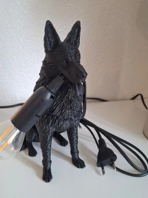 Bordslampa schäferhund  - Unik bordslampa formad som en schäferhund i svart plast. Lampan har en glödlampa som hålls i hundens mun och svart sladd med stickkontakt. Perfekt som cool inredningsdetalj för hundälskare eller dig som vill ha något annorlunda.