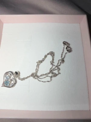Halsband med hjärthänge i silver - Snyggt halsband i silverfärg med ett hjärthänge. Hänget har en glittrande blå sten och en klar sten, vilket ger en elegant och romantisk look. Kedjan är tunn och tvinnad, perfekt för att lyfta din outfit.