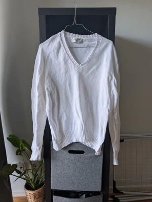 Vit stickad tröja med v-ringning - Klassisk vit stickad tröja med v-ringad hals och ribbade muddar vid ärmslut och nederkant. Tröjan har lång ärm och är tillverkad i ett mjukt, tunt stickat material som passar perfekt till lager på lager. Enkel och stilren design.