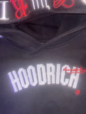 Svart med röda detaljer hoodie från hoodrich 160-165 - Säljer en svart hoodie från Hoodrich med stor vit broderad logga på bröstet och röda detaljer. Klassisk huva och mjukt material som känns skönt mot huden. Perfekt för dig som gillar streetwear och vill ha en stilren look.