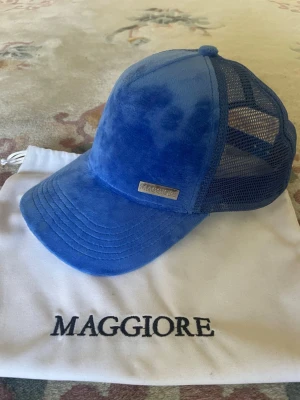 Blå keps i sammet från Maggiore - Snygg blå keps från Maggiore i sammet med mesh på sidorna och bak. Kepsen har böjd skärm och metallogo framtill. 