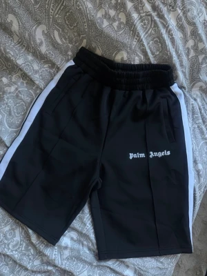 Palm Angels svarta shorts med vita ränder - Svarta shorts från Palm Angels med vita sidoränder och elastisk midja. Klassisk logga broderad framtill och sportig look. Perfekta för en avslappnad streetwear-stil. Materialet känns mjukt och följsamt, troligen polyester.