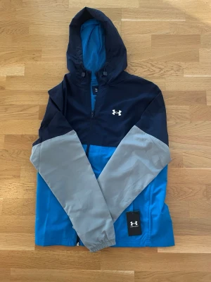 Blå och grå vindjacka från Under Armour - Snygg vindjacka från Under Armour i blått, mörkblått och grått. Jackan har huva med dragsko, hel dragkedja och Under Armour-logga på bröstet. Perfekt för blåsiga dagar och sportiga outfits.