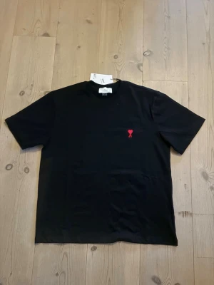Svart t-shirt från Ami Paris - Svart t-shirt från Ami Paris med klassisk passform och korta ärmar. Tillverkad i mjuk bomull och har en liten röd broderad logga på bröstet. Perfekt basplagg med stilren design och rund halsringning.