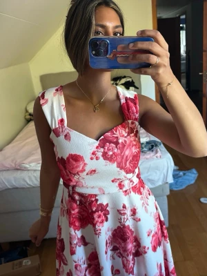 Vit klänning med röda blommor - Superfin vit klänning med stora röda blommor och breda axelband. Klänningen har en markerad midja och en utsvängd kjol som ger en feminin siluett. Perfekt för sommarens alla tillfällen! Materialet känns lätt och bekvämt. Säljer då den är för stor för mig och kommer inte till användning!