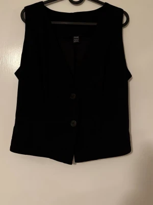 Svart väst från SHEIN med knappar - Stilren svart väst från SHEIN med två stora knappar framtill och v-ringad hals. Västen har en klassisk rak passform och är tillverkad i ett mjukt syntetmaterial. Perfekt att styla över en skjorta eller t-shirt för en trendig look.