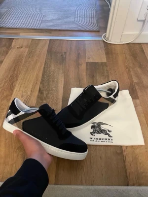 Burberry svarta sneakers med rutigt mönster - Snygga svarta sneakers från Burberry med klassiskt rutigt mönster på hälen. Skorna har vita sulor, svarta snören och detaljer i mocka och textil. Perfekta för dig som vill ha en stilren och lyxig look med en touch av ikoniskt brittiskt mönster. Storlek 43 passar 42