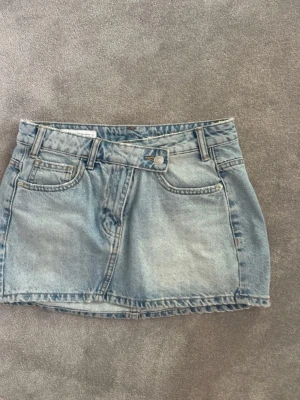 Ljusblå jeanskjol med fickor - Trendig ljusblå jeanskjol i kort modell med klassiska fem fickor, bälteshällor och knappstängning fram. Perfekt för dig som gillar en avslappnad och cool stil. Passar till allt från sneakers till boots.