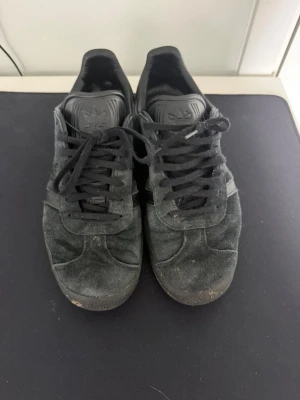Svarta Adidas sneakers i mocka - Svarta sneakers från Adidas med klassisk låg siluett och ovandel i mocka. Skorna har svarta skosnören, diskret Adidas-logga på plösen och sula i gummi. Perfekta för en stilren och avslappnad look.