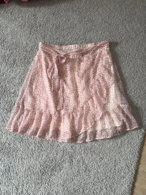 Rosa omlottkjol med volang - Söt rosa omlottkjol med små blommigt mönster och volangkant. Kjolen har knytband i midjan och är tillverkad i ett lätt och luftigt material, 