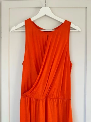 Korallorange långklänning / maxiklänning med omlottdetalj - Snygg  långklänning med omlottdetalj framtill och ärmlös design. Klänningen har en avslappnad passform och är tillverkad i ett mjukt, lätt material som passar perfekt för varma dagar. Märklapp och storlekslapp saknas, är dock från Gina tricot. Mått: Längd: ca 136 cm Midja (outsträckt): 38 cm. Slitsarna på bägge sidor är igensydda en liten bit, går ungefär till mitt på vaden just nu. Mycket fint skick, använd max 1-2ggr. 