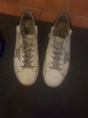 Vita Golden Goose sneakers med stjärna - Säljer ett par vita Golden Goose sneakers i storlek 44 passar även 43 med grå stjärna på sidan och grå snörning. Skorna har lågt skaft och detaljer i beige och svart på hälen. Klassisk design i läder som passar till det mesta. Hör av er vid frågor😊