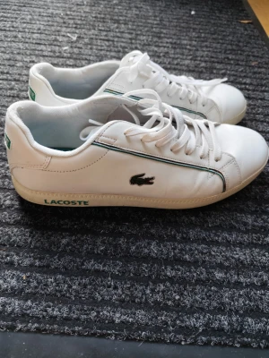 Vita Lacoste sneakers med gröna detaljer - Fräscha vita sneakers från Lacoste med klassisk krokodillogga på sidan och gröna detaljer längs kanten. Skorna har lågt skaft, vita snören och diskret Lacoste-tryck på sulan. Perfekta för en clean och sportig stil.