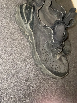Svarta Nike react skor - Svarta Nike react skor med mesh- och syntetmaterial, chunky sula och klassisk Air Max-dämpning. Skorna har svarta skosnören och detaljer samt en sportig vibe som passar streetwear-stilen.