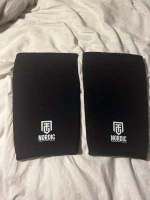 Svarta knäskydd från Nordic Training Gear - Ett par svarta knäskydd från Nordic Training Gear, perfekta för styrketräning eller crossfit. De är tillverkade i ett stretchigt och slitstarkt material med vit logga framtill. Sitter tajt runt knäna för extra stöd under tunga lyft.