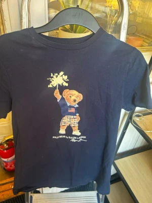 Mörkblå Polo Bear t-shirt Ralph Lauren - Mörkblå t-shirt från Ralph Lauren med ikoniska Polo Bear-trycket framtill. Björnen har en amerikansk flaggtröja och rutiga byxor. Klassisk rund hals och korta ärmar. Perfekt för dig som gillar streetwear och vill ha något stilrent men lekfullt.