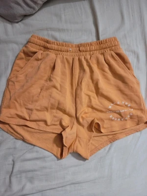 Persikofärgade mjukisshorts - Säljer ett par persikofärgade mjukisshorts med resår i midjan och fickor på sidorna. Tryck i vitt på ena benet med texten 'MAKE NEW MEMORIES EVERYDAY'. Perfekta för chill dagar eller träning.