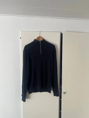 Mörkblå stickad tröja med halvzip - Mörkblå stickad tröja med ståkrage och halv dragkedja framtill. Tröjan har ribbstickat mönster och långa ärmar. Perfekt för lager-på-lager och enkel att matcha med jeans eller chinos. Hör av er vid frågor!