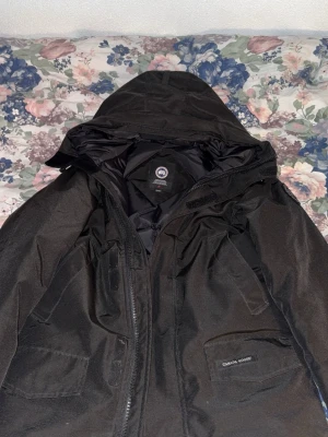Svart dunjacka från Canada Goose (Black Label) - Säljer en svart dunjacka från Canada Goose med huva och flera praktiska fickor. Jackan har diskret logotyp på ärmen och är fodrad för att hålla dig varm under kalla vinterdagar. Perfekt för dig som vill ha både stil och funktion.