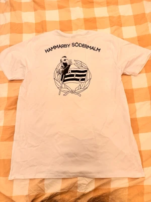 Vit Hammarby Södermalm t-shirt - Vit t-shirt med svart tryck på ryggen som visar en person, flagga och texten 'Hammarby Södermalm'. Klassisk passform och rund hals. Perfekt för dig som vill visa ditt stöd för Hammarby.